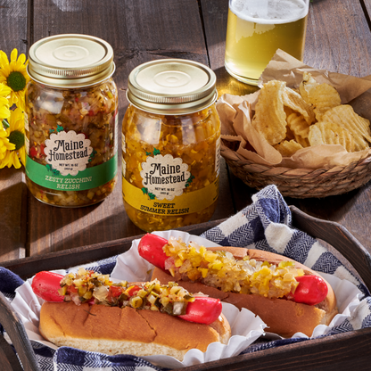 Two jars of Maine Homestead pickled relish displayed on a rustic wooden table, with two hot dogs topped with relish in the foreground and a basket of potato chips and a glass of beer nearby.