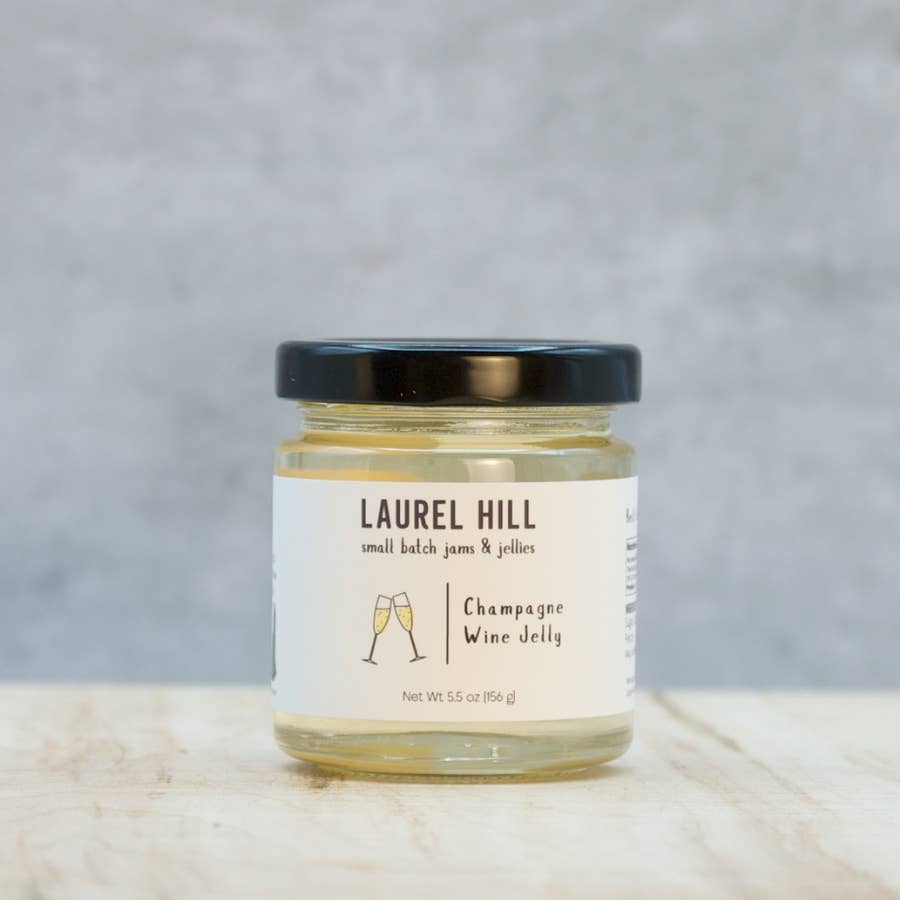 A jar of Laurel Hill Champagne Wine Jelly sits on a light wooden surface against a textured gray background. The clear glass jar shows the pale yellow jelly inside, topped with a black lid and featuring a white label with black text and a small champagne glass illustration.