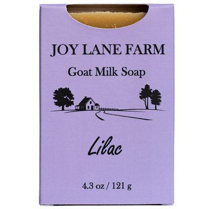 A rectangular lavender soap box labeled 'JOY LANE FARM Goat Milk Soap' with 'Lilac' printed near the bottom and a small black farm illustration. A curved cutout at the top reveals the golden soap bar inside.