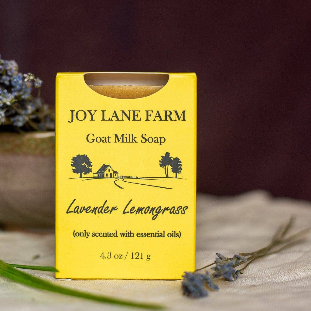 A yellow box of Joy Lane Farm Goat Milk Soap, scented with Lavender Lemongrass, displayed on a textured fabric surface with dried lavender sprigs and a rustic pot in the background.
