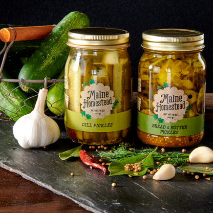 Two jars of pickles with 'Maine Homestead' labels on a dark surface with cucumbers and garlic.