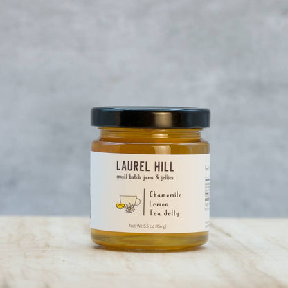 A jar of Laurel Hill Chamomile Lemon Tea Jelly on a light marble surface with a textured gray background. The product is in a clear glass jar filled with golden-yellow jelly, featuring a cream-colored label and a black lid.