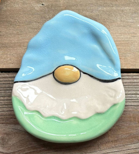A ceramic gnome head wall decor piece with a light blue hat, cream beard, mint green base, and a small yellow nose, displayed on a rustic wooden surface.