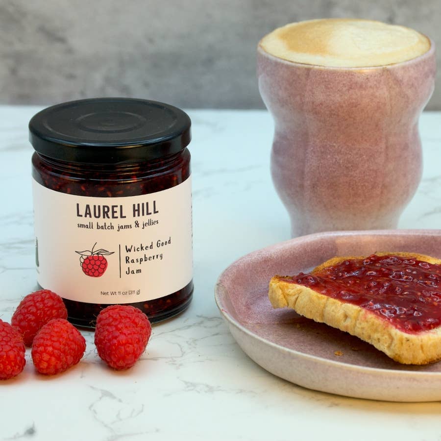 Jar of Laurel Hill raspberry jam with a spread on toast and raspberries on a marble surface.