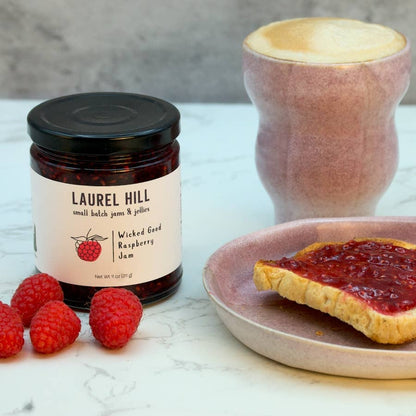 Jar of Laurel Hill raspberry jam with a spread on toast and raspberries on a marble surface.