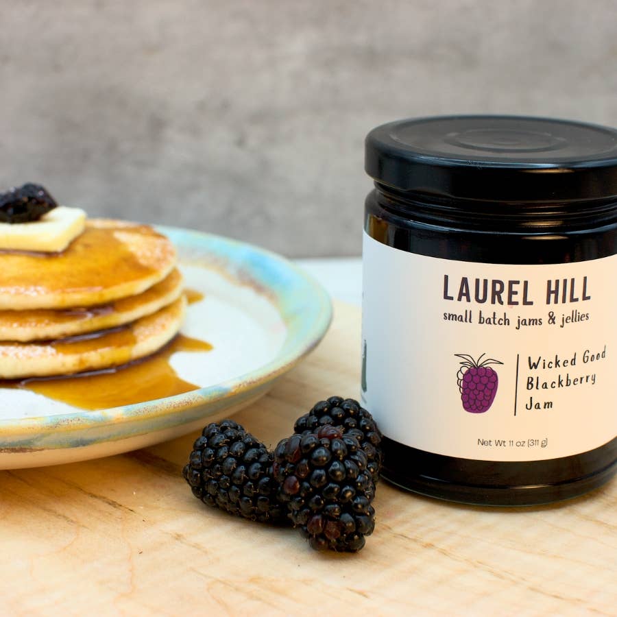 Jar of Laurel Hill Wicked Good Blackberry Jam with pancakes and blackberries on a wooden surface