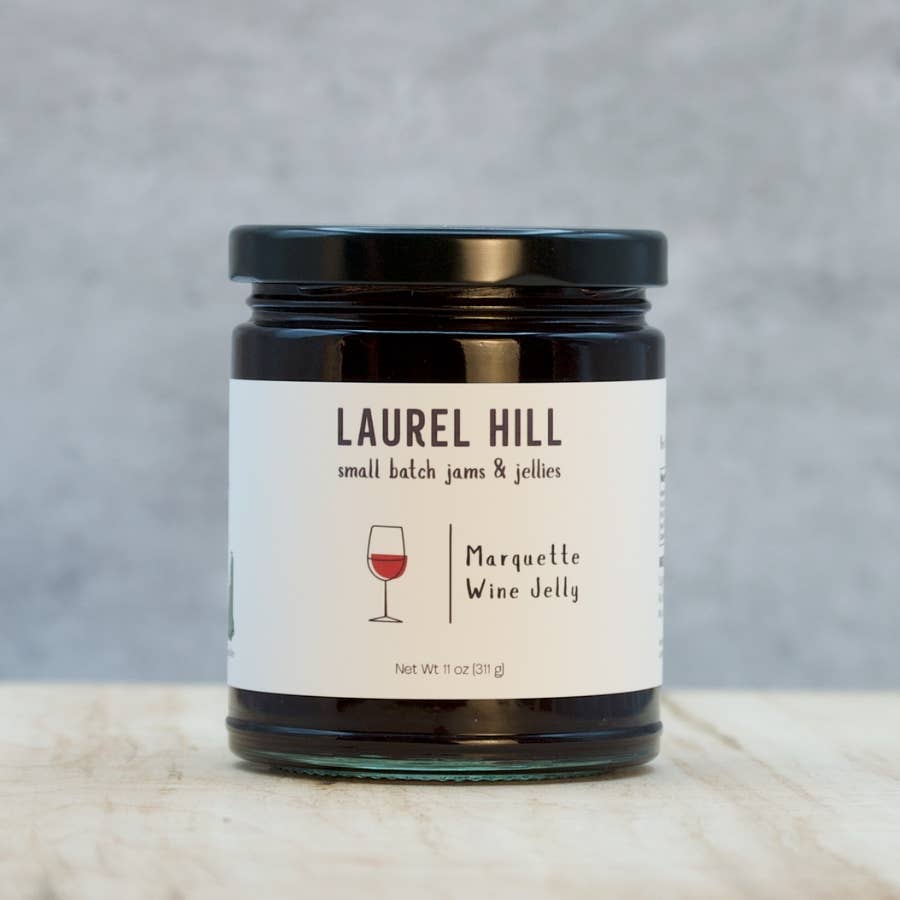 A glass jar of Laurel Hill Small Batch Jams & Jellies, specifically Marquette Wine Jelly, with a white label and black lid, set on a light marble surface against a textured gray background.