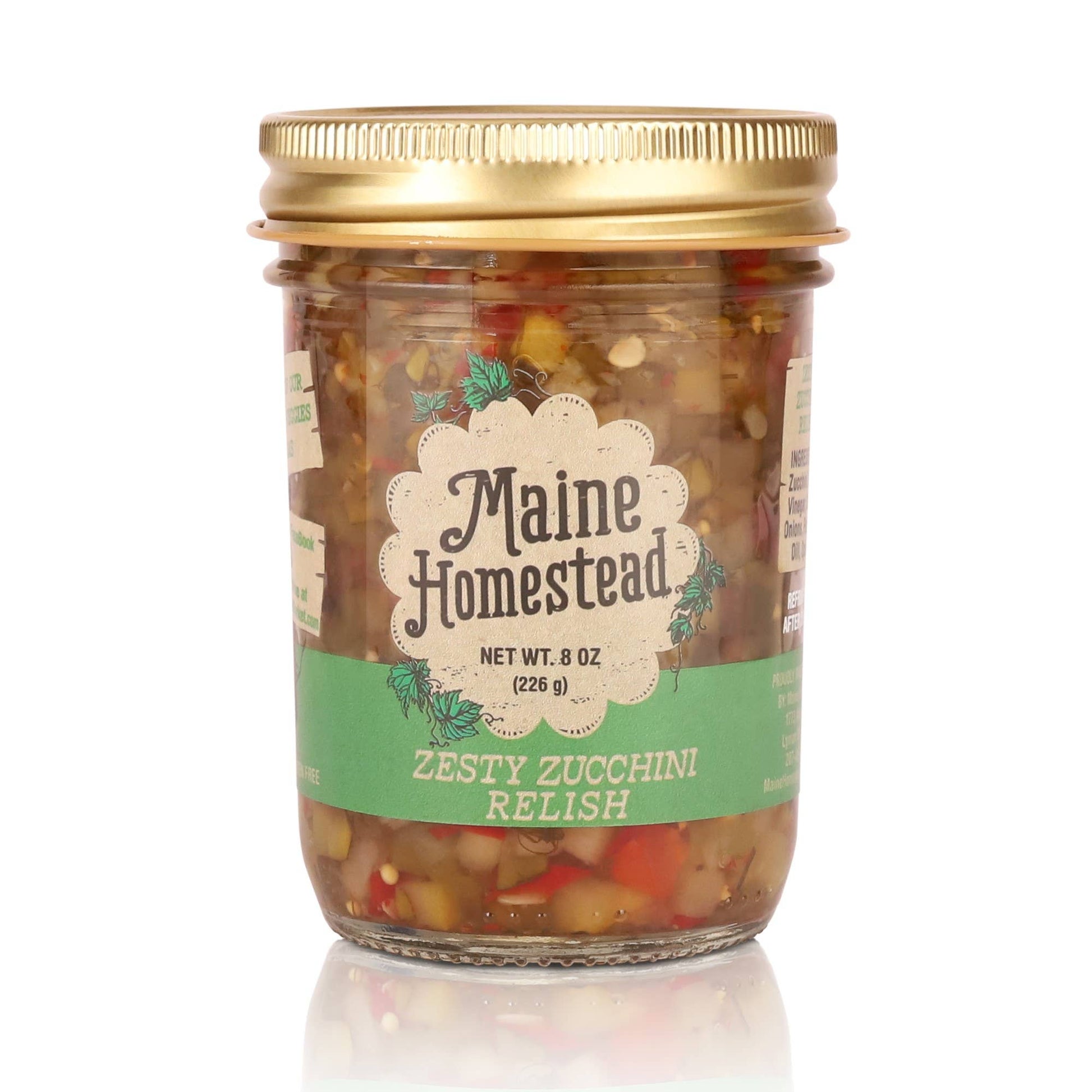 A glass jar of Maine Homestead Zesty Zucchini Relish with a gold metal lid and a green and white label, set against a plain white background with a soft reflection below.