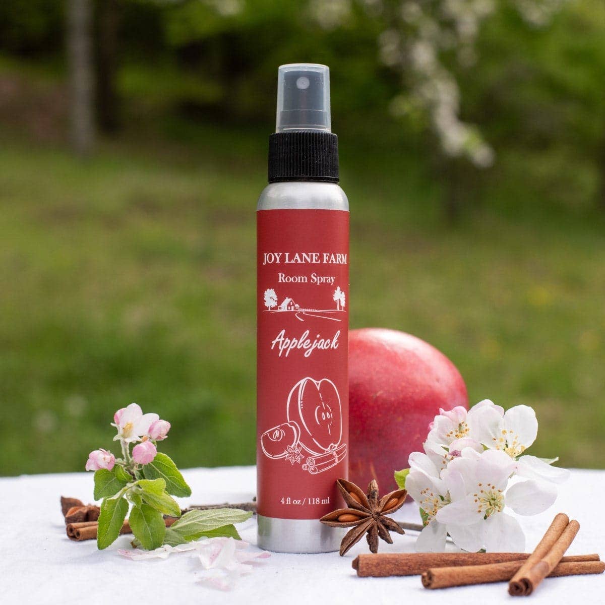 A bottle of Joy Lane Farm Applejack Room Spray stands on a white cloth outdoors, surrounded by apple blossoms, cinnamon sticks, star anise, and a red apple, with a blurred green garden background.