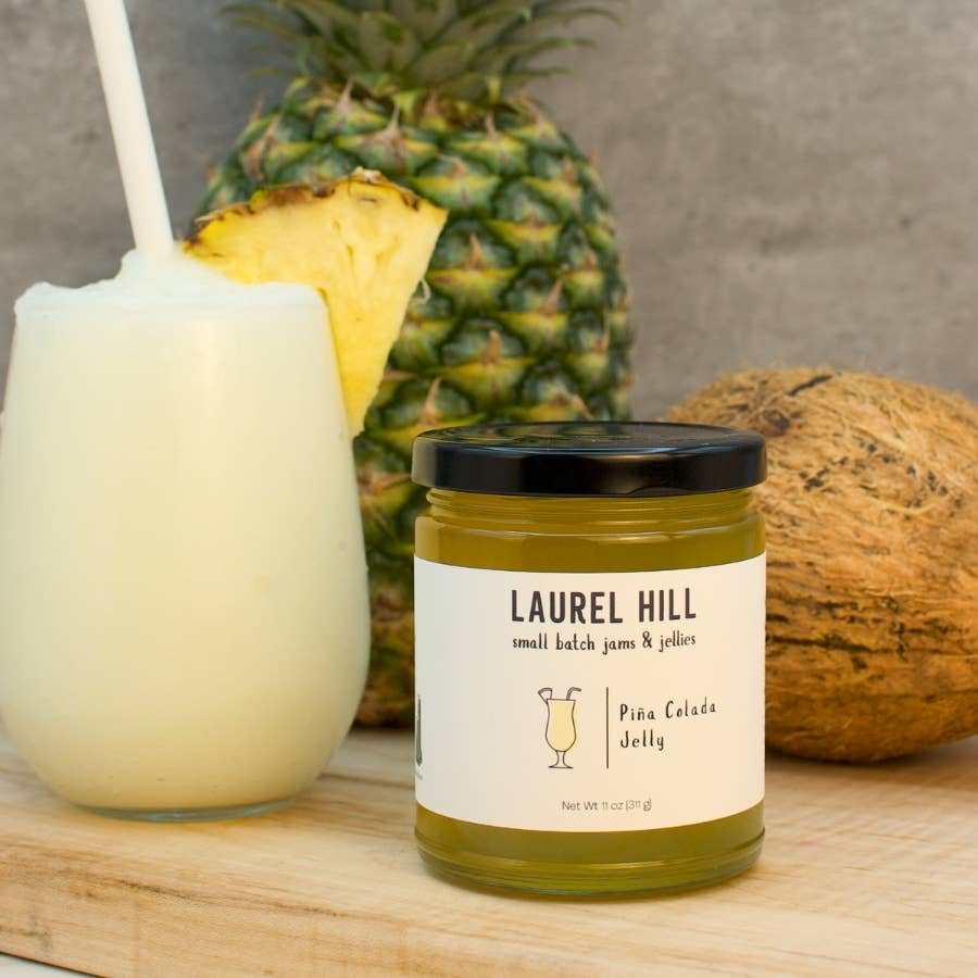 A jar of Laurel Hill Pina Colada Jelly on a wooden board, styled with a pineapple, a coconut, and a tropical drink with a straw.