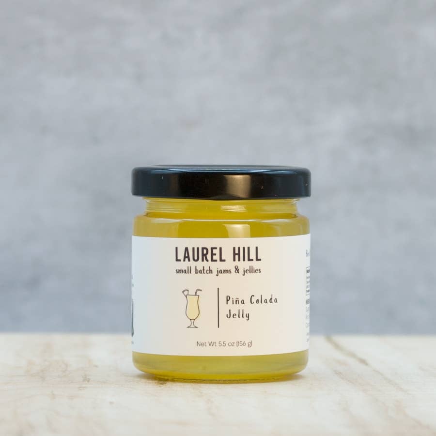 A jar of Laurel Hill Pina Colada Jelly on a light marble surface with a textured gray background. The clear glass jar shows the yellow jelly inside, has a black lid, and a white label with black text.