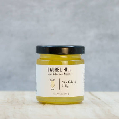 A jar of Laurel Hill Pina Colada Jelly on a light marble surface with a textured gray background. The clear glass jar shows the yellow jelly inside, has a black lid, and a white label with black text.