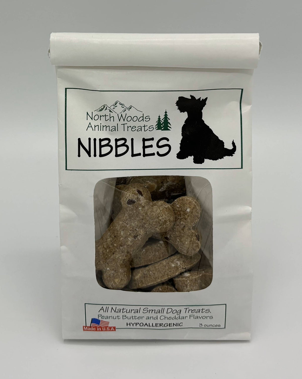 A white stand-up pouch of North Woods Animal Treats Nibbles dog treats, with a clear window showing the brown bone-shaped treats inside. The packaging features a black dog silhouette and text indicating all natural small dog treats in peanut butter and cheddar flavors.