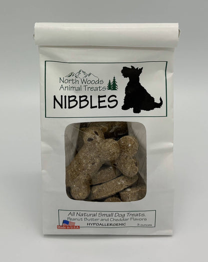 A white stand-up pouch of North Woods Animal Treats Nibbles dog treats, with a clear window showing the brown bone-shaped treats inside. The packaging features a black dog silhouette and text indicating all natural small dog treats in peanut butter and cheddar flavors.