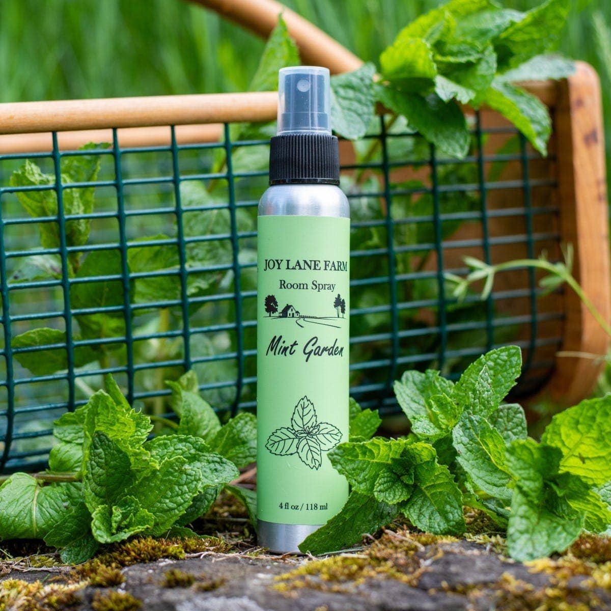 A room spray bottle labeled 'Joy Lane Farm Mint Garden' stands in front of a green wire garden basket filled with mint leaves, with real mint foliage and moss around the base.