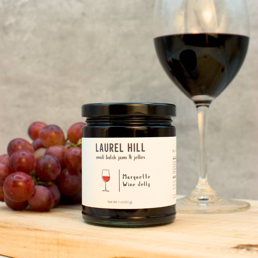 A jar of Laurel Hill Marquette Wine Jelly sits on a wooden board with a glass of red wine and red grapes in the background against a textured gray wall.