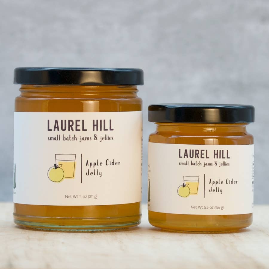 Two glass jars of Laurel Hill Apple Cider Jelly, one larger and one smaller, with cream labels and black lids, set on a light marble surface against a gray background.