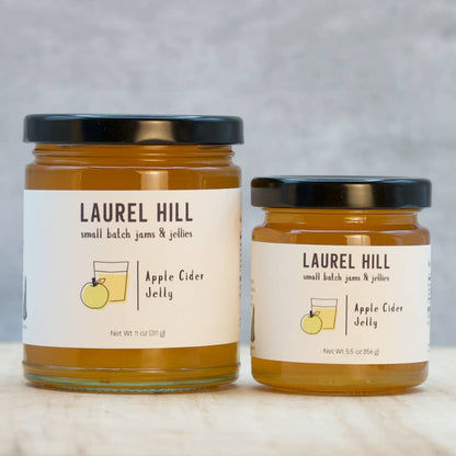 Two glass jars of Laurel Hill Apple Cider Jelly, one larger and one smaller, with cream labels and black lids, set on a light marble surface against a gray background.