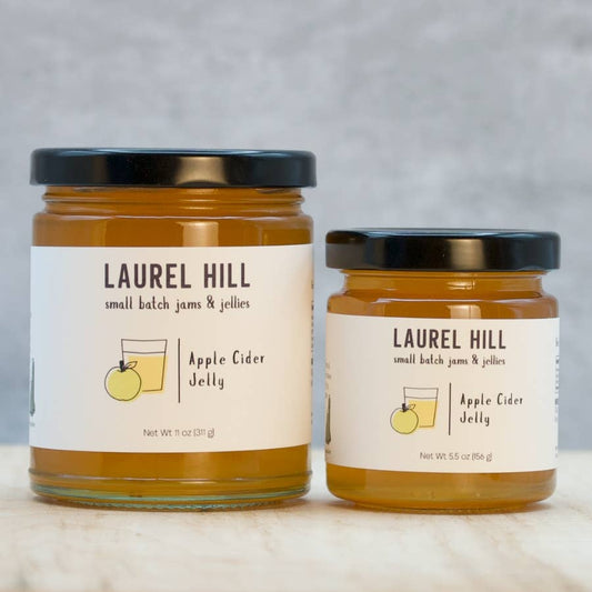 Two glass jars of Laurel Hill Apple Cider Jelly, one larger and one smaller, with cream labels and black lids, set on a light marble surface against a gray background.