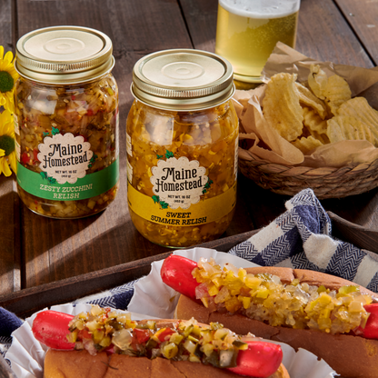 Two jars of Maine Homestead relish on a rustic wooden table, with a hot dog topped with relish in the foreground and potato chips and a glass of beer in the background.