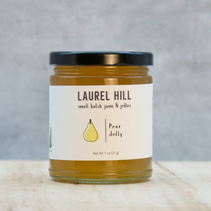 A glass jar of Laurel Hill Pear Jelly with a black lid and a white label, set on a light wooden surface against a textured gray background.