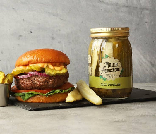 Hamburger with pickles and a jar of Maine Homestead Dill Pickles on a gray surface.