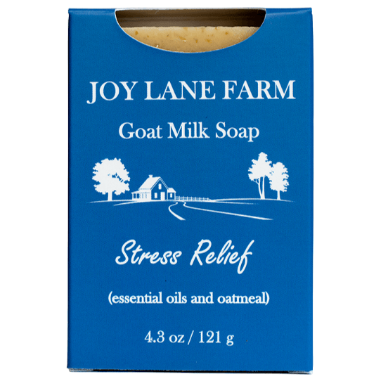 A rectangular blue box of Joy Lane Farm Goat Milk Soap, labeled 'Stress Relief' with white text and a small white farm illustration. A small piece of beige soap is visible at the top through a die-cut window, on a plain white background.