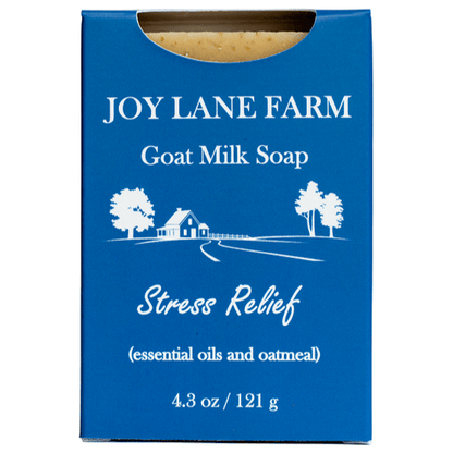 A rectangular blue box of Joy Lane Farm Goat Milk Soap, labeled 'Stress Relief' with white text and a small white farm illustration. A small piece of beige soap is visible at the top through a die-cut window, on a plain white background.