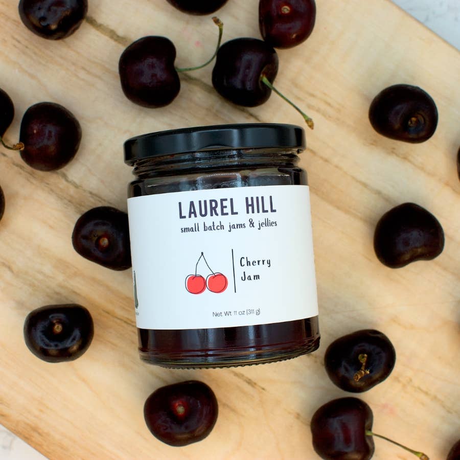 A glass jar of Laurel Hill Cherry Jam with a black lid and white label, surrounded by fresh dark cherries on a light wood surface.