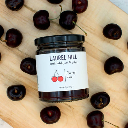 A glass jar of Laurel Hill Cherry Jam with a black lid and white label, surrounded by fresh dark cherries on a light wood surface.