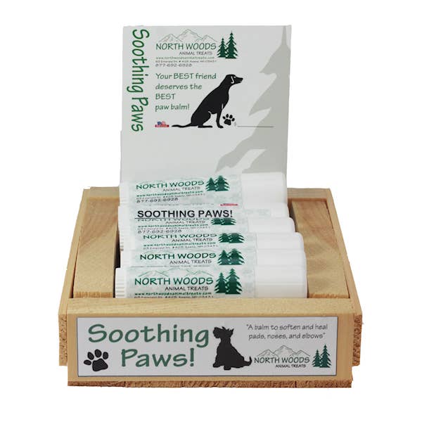 A wooden display tray holding several packets of North Woods Animal Treats Soothing Paws pet balm, with one packet standing upright behind the others on a white background.