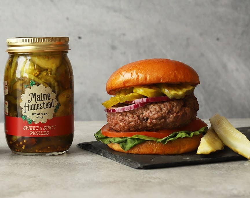 Burger with pickles and a jar of Maine Homestead pickles on a gray surface.