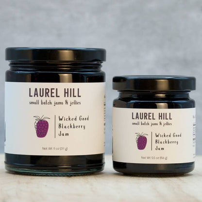Two jars of Laurel Hill Wicked Good Blackberry Jam on a marble surface with a gray background.