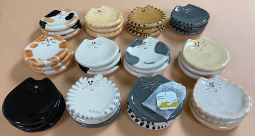 A collection of ceramic cat-shaped dishware and small containers in assorted colors, including black, white, gray, beige, and orange, arranged on a solid peach-colored background.