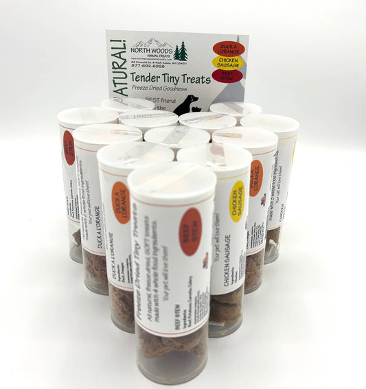 A group of clear plastic treat jars filled with small brown treats, arranged in front of a branded North Woods Pet Supplies card on a white studio background.