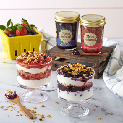 Two layered dessert parfaits with granola and berries, accompanied by jars of Maine Homestead jam on a marble surface.