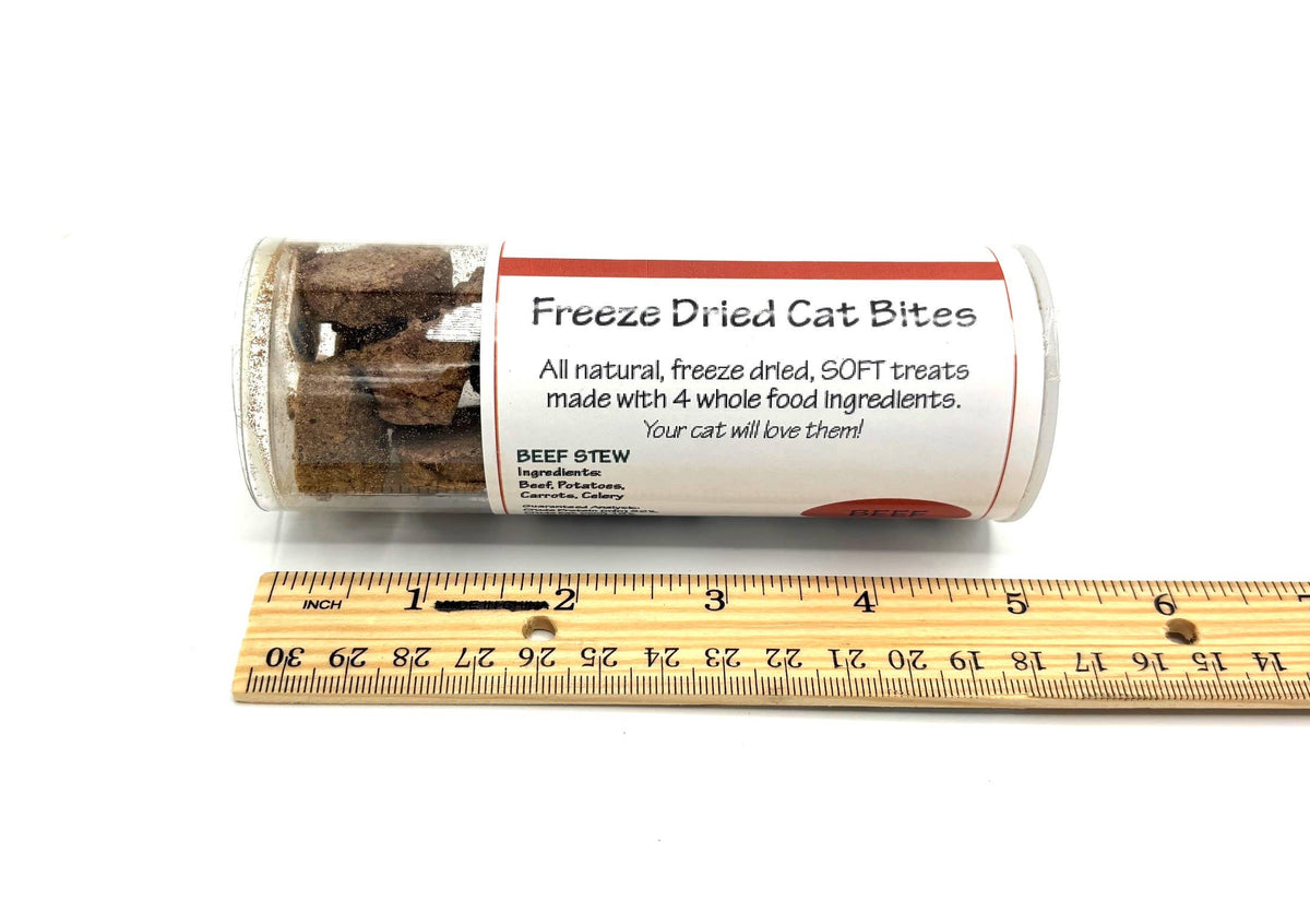 A clear plastic tube of freeze dried cat bites treats with a white label, shown next to a wooden ruler on a white background. The label reads 'Freeze Dried Cat Bites' and 'BEEF STEW'.