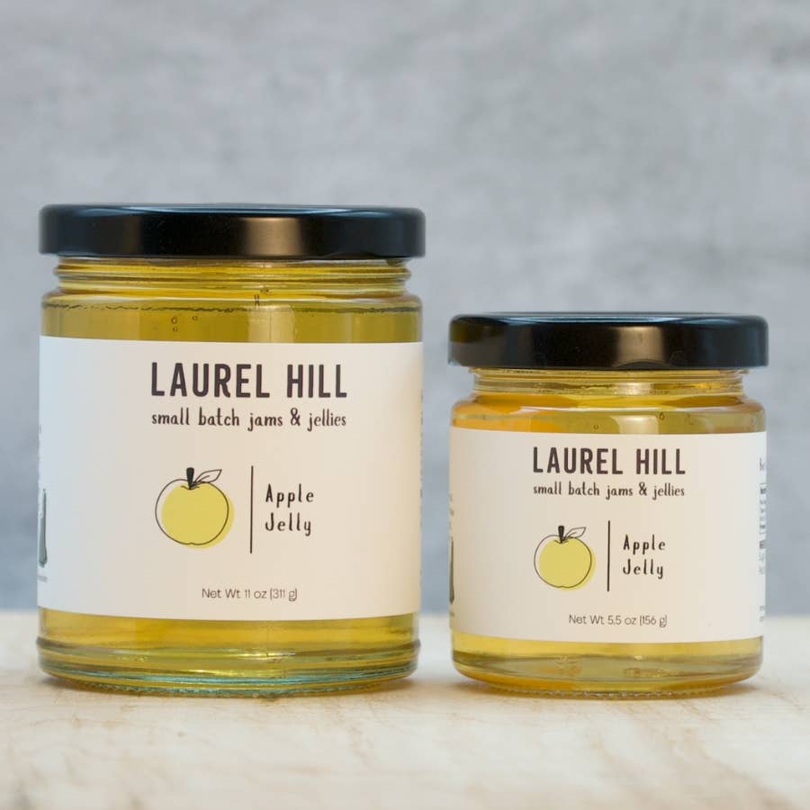 Two glass jars of Laurel Hill Apple Jelly, one larger 11 oz jar and one smaller 5.5 oz jar, both filled with yellow apple jelly and featuring white labels with black text and a yellow apple illustration, set on a light marble surface against a gray background.