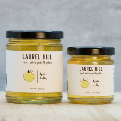 Two glass jars of Laurel Hill Apple Jelly, one larger 11 oz jar and one smaller 5.5 oz jar, both filled with yellow apple jelly and featuring white labels with black text and a yellow apple illustration, set on a light marble surface against a gray background.