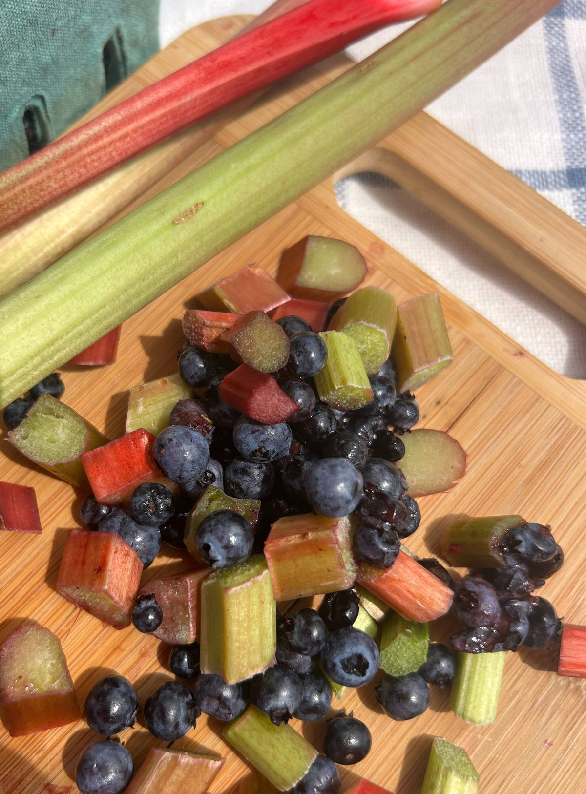 Chopped rhubarb and blueberries on a wooden cutting board, with whole rhubarb stalks placed behind them.