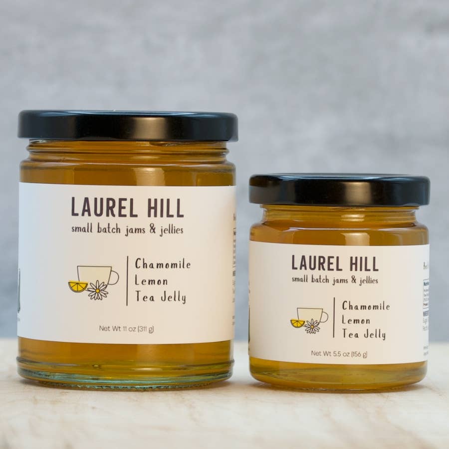 Two glass jars of Laurel Hill Small Batch Jams & Jellies, one larger 9 oz jar and one smaller 5.5 oz jar, both filled with chamomile lemon tea jelly and topped with black lids, set on a light marble surface against a gray textured background.