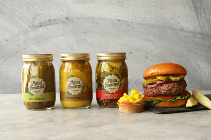 Three jars of Maine Homestyle pickles with a burger and condiments on a gray surface.