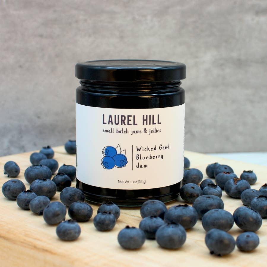 Jar of Laurel Hill Wicked Good Blueberry Jam surrounded by blueberries on a wooden surface.