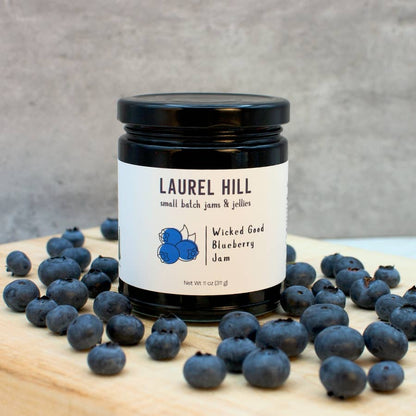 Jar of Laurel Hill Wicked Good Blueberry Jam surrounded by blueberries on a wooden surface.