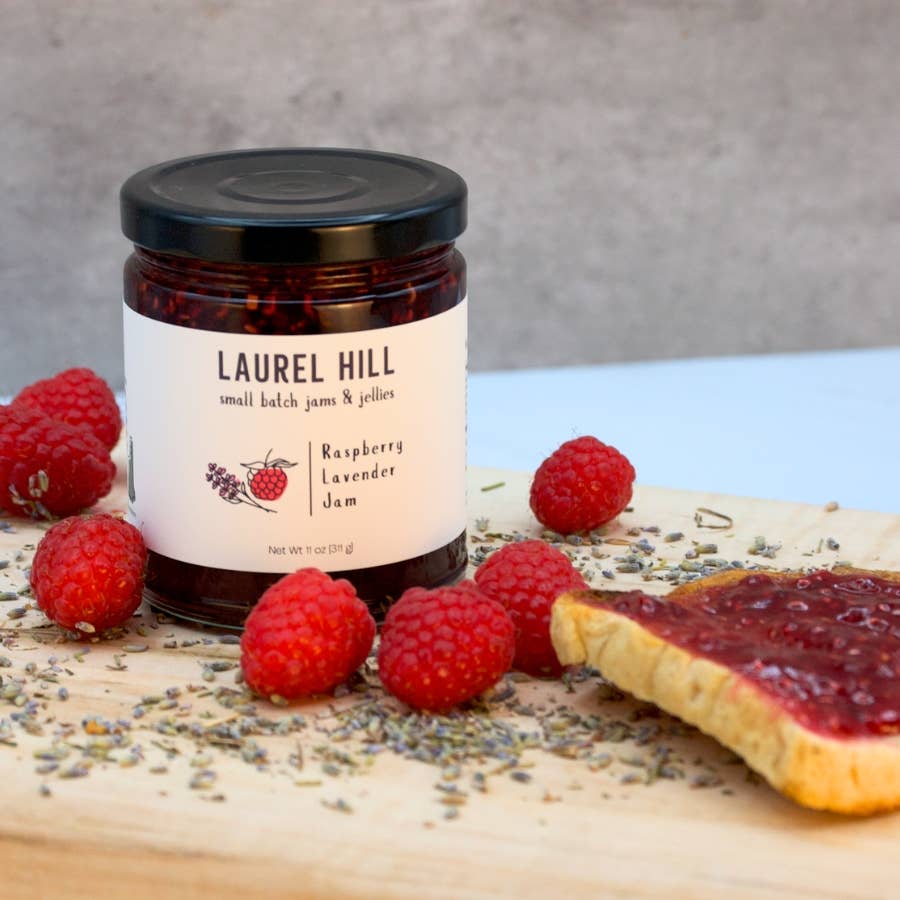 Jar of Laurel Hill raspberry lavender jam with raspberries and a slice of bread on a wooden board.