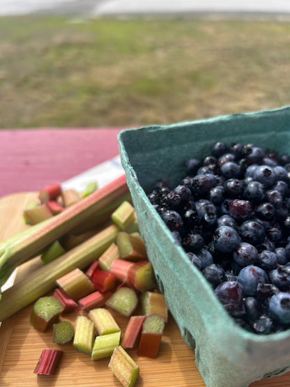 A green paperboard berry carton filled with blueberries on a wooden cutting board next to chopped rhubarb stalks, with an outdoor blurred background.