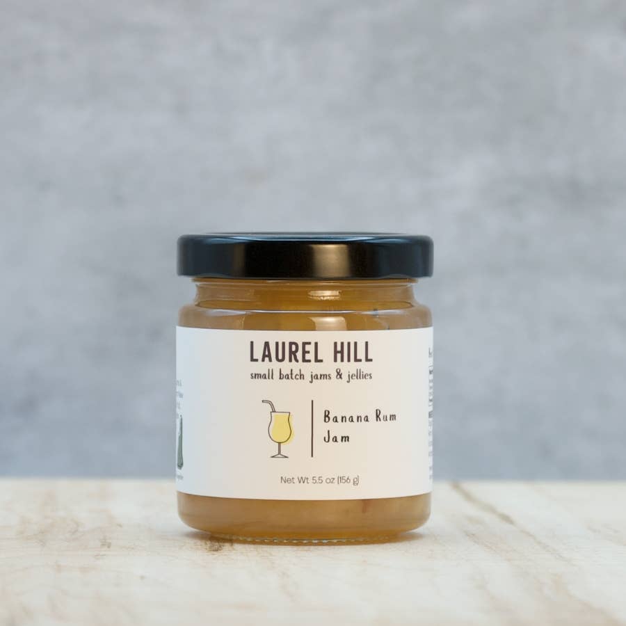 A jar of Laurel Hill Small Batch Jams & Jellies Banana Ram Jam with a black lid and white label, set on a light wooden surface against a gray textured background.