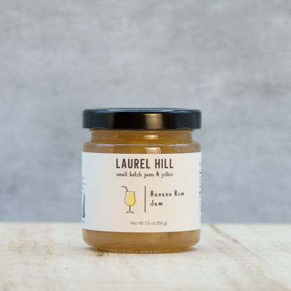 A jar of Laurel Hill Small Batch Jams & Jellies Banana Ram Jam with a black lid and white label, set on a light wooden surface against a gray textured background.