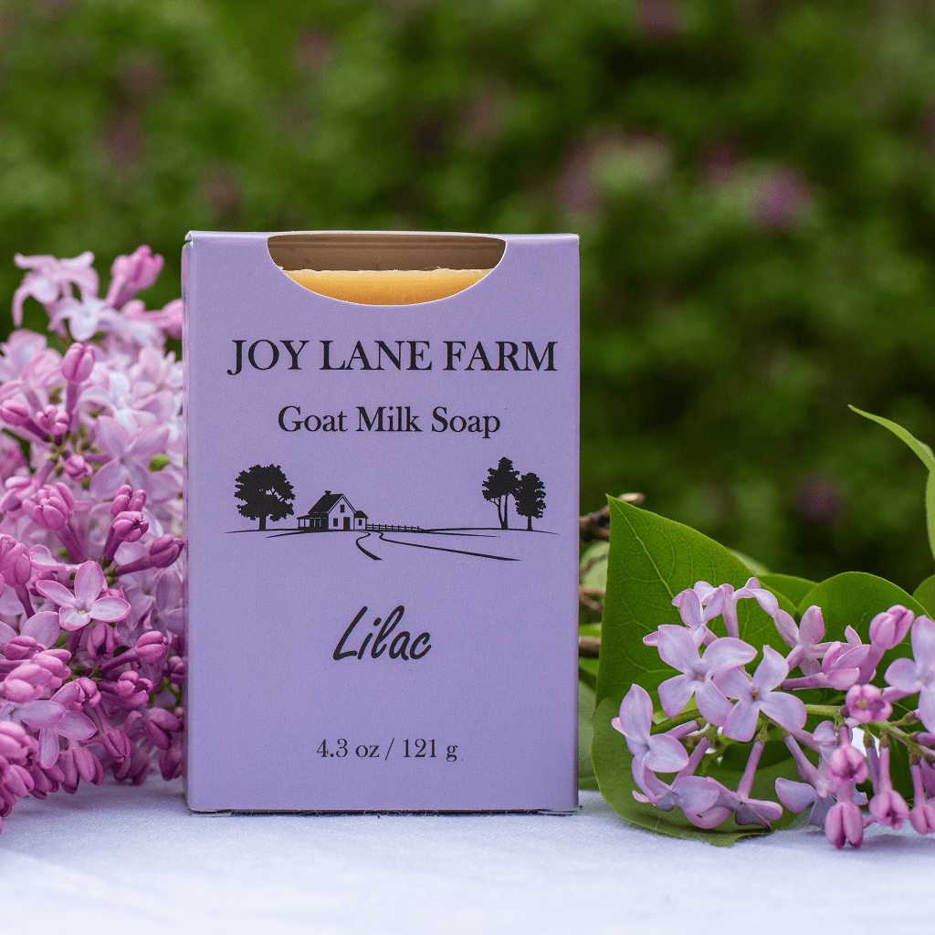 A lavender soap bar in a matching box labeled Joy Lane Farm Goat Milk Soap, Lilac, displayed outdoors on a white surface with pink flowers and a blurred green garden background.