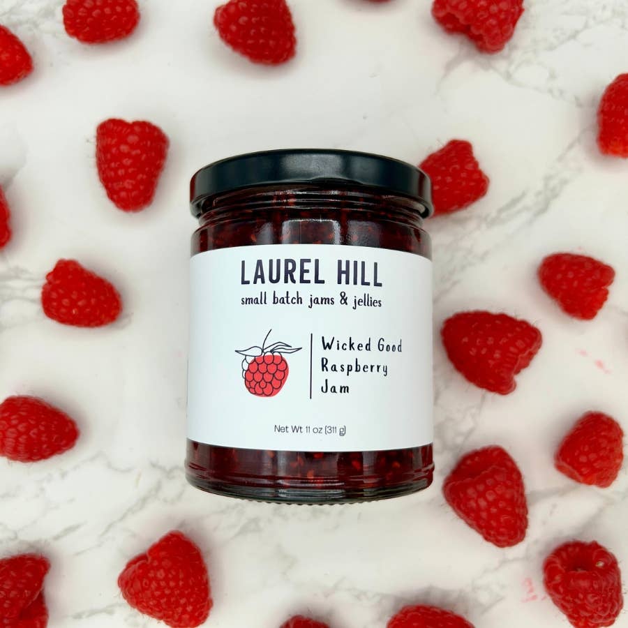 Jar of Laurel Hill raspberry jam surrounded by raspberries on a white background